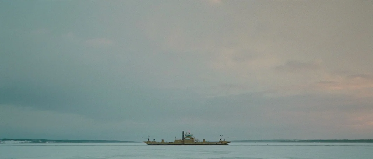 The American (2010) movie still: establishing — A wide, desolate landscape shows a yellow ferry crossing a vast expanse of ice. The flat horizon is…