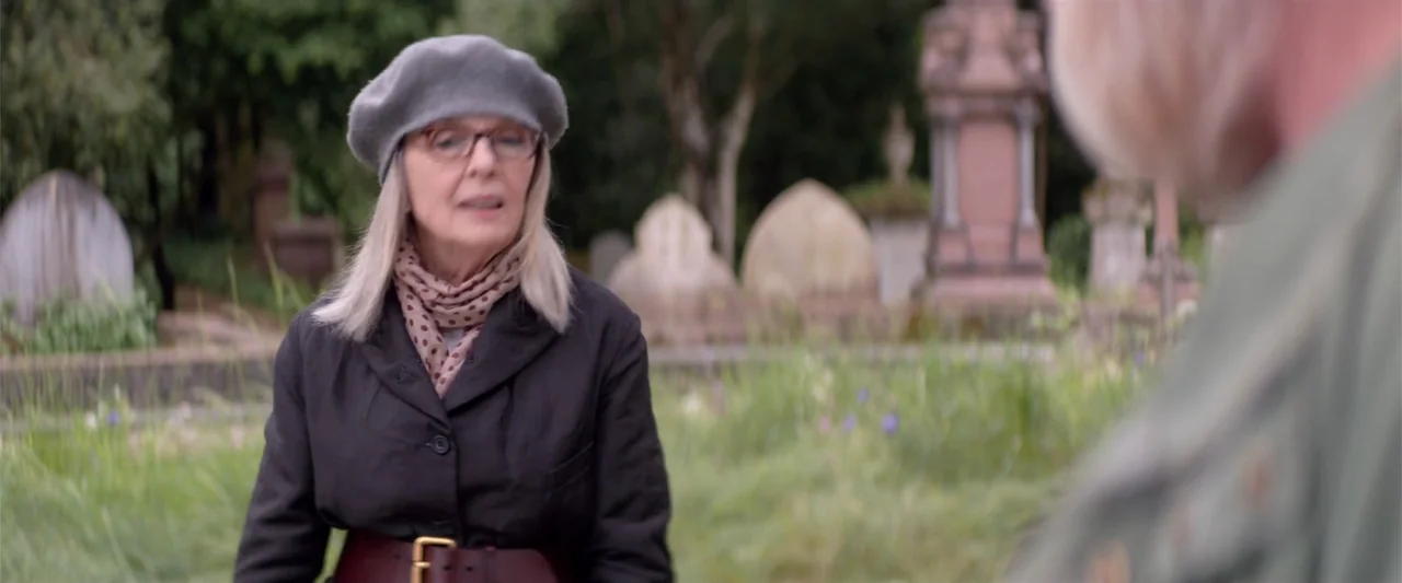 Hampstead (2017) movie still: medium — A quiet cemetery with aged stone monuments and tall green grass. An older woman in a dark jacket stands…