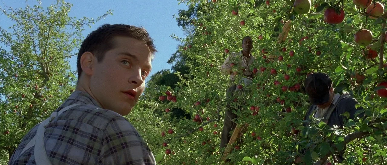 The Cider House Rules movie still: medium — An apple orchard is filled with trees laden with ripe red fruit. A young man in a plaid shirt turns his…
