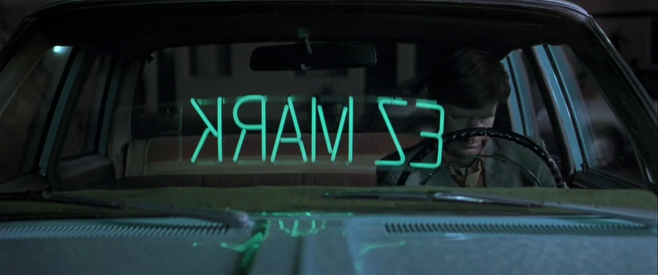 The Cooler (2003) movie still: medium — Inside a car cabin at night with a neon sign visible through the windshield. A man sits in the driver seat…