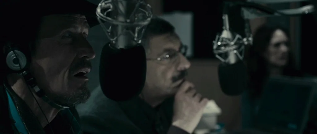 Pontypool (2008) movie still: medium — Two men sit side-by-side in a dim, modern audio recording booth with soundproofing equipment. They speak into…