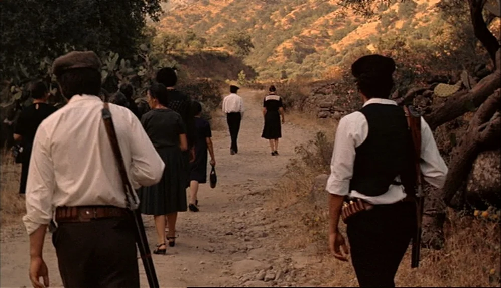 The Godfather (1972) movie still: fullbody — A group of people walks away along a dusty rural path lined by scrub vegetation. Hilly terrain rises in…