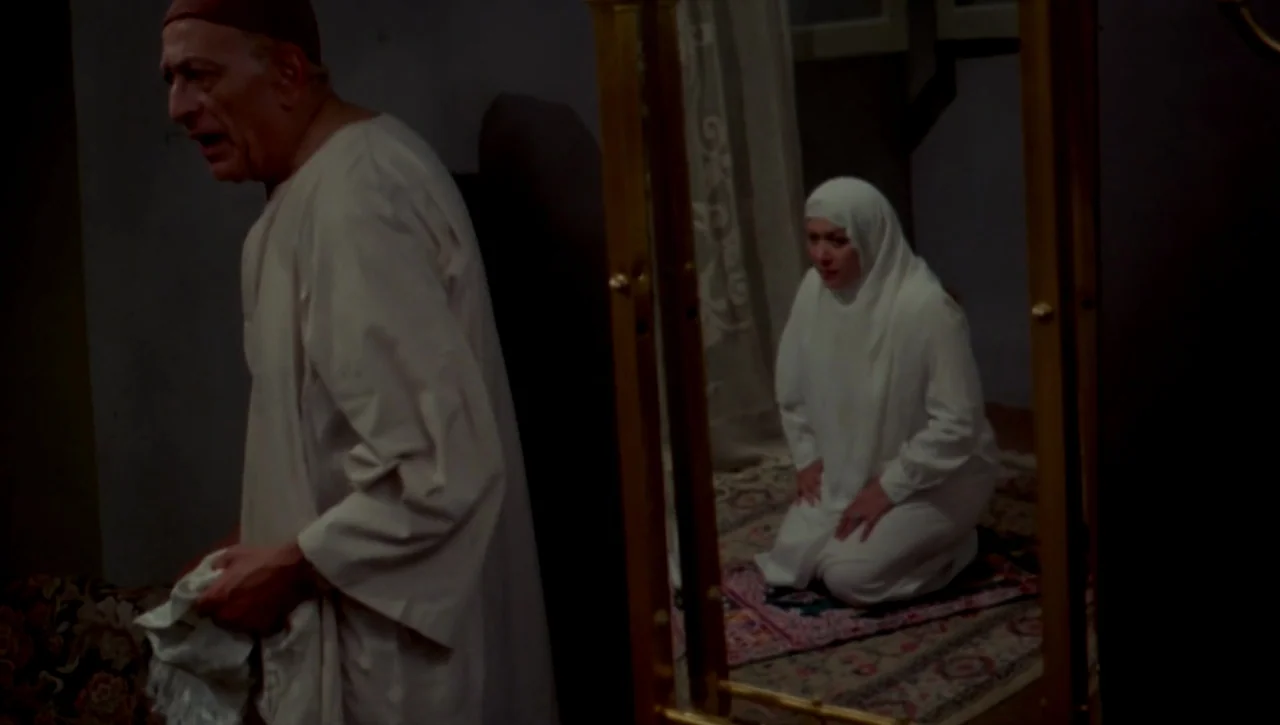 The Return of the Prodigal Son (1978) movie still: medium — A dimly lit room with a man standing beside a large floor mirror. A woman wearing white…