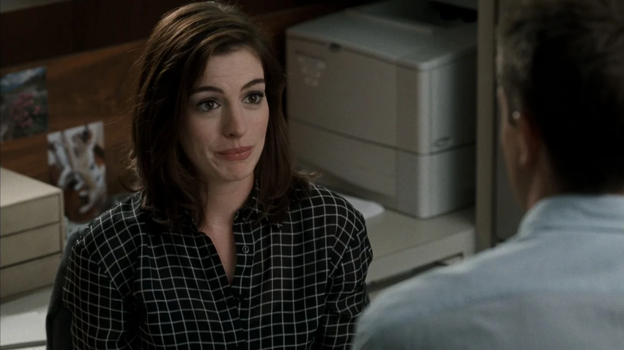F Valentine's Day (2026) movie still: medium — A workspace features a desk with a white printer and wooden partition walls. A woman in a dark…