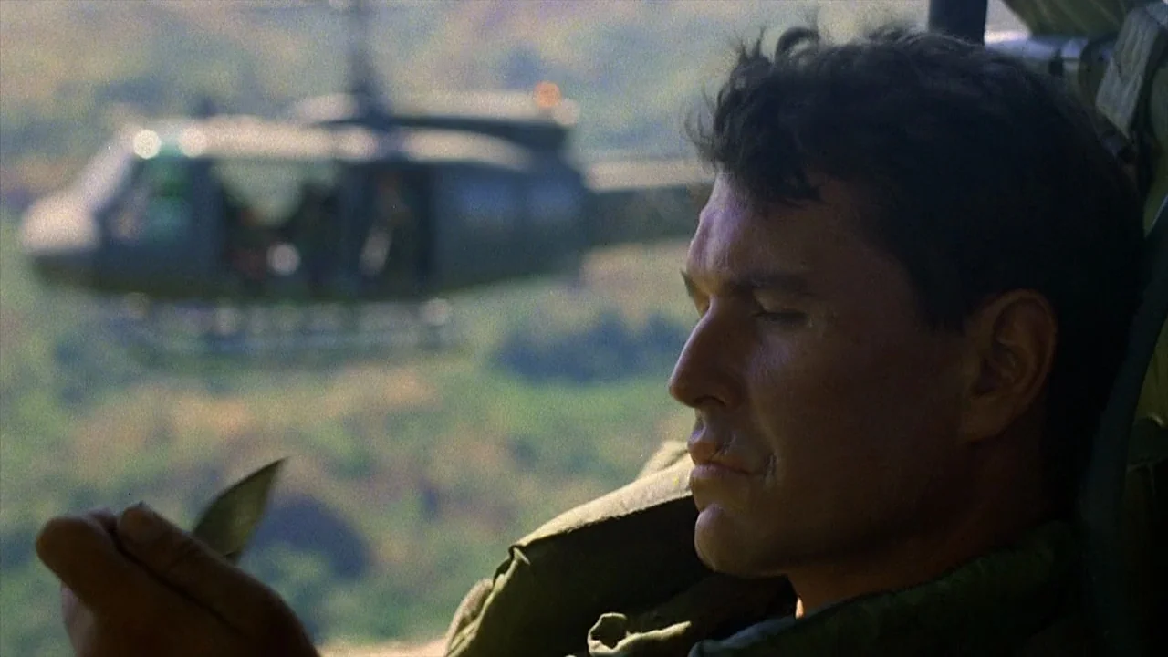 Platoon (1986) movie still: medium — A man rides in an open military helicopter flying above a dense green canopy. He sits in profile looking down…