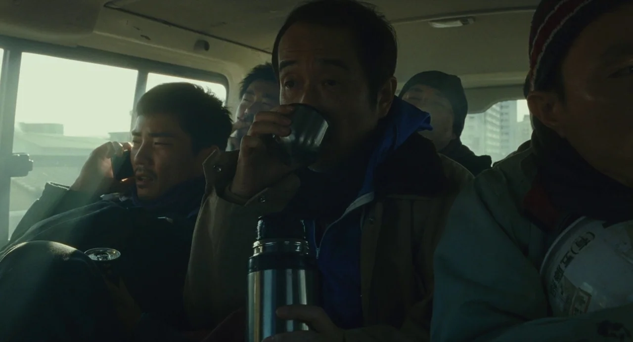 Shoplifters (2018) movie still: medium — A group of men sit inside the back of a van during the day. The primary subject drinks from a metallic…