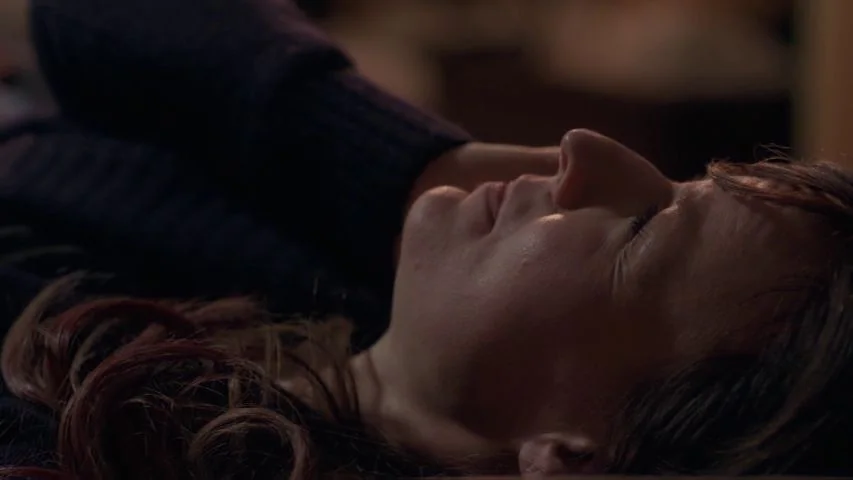 Touchy Feely (2013) movie still: closeup — A woman rests her head on a dark surface in a dimly lit room. She wears a navy sweater and keeps her eyes…