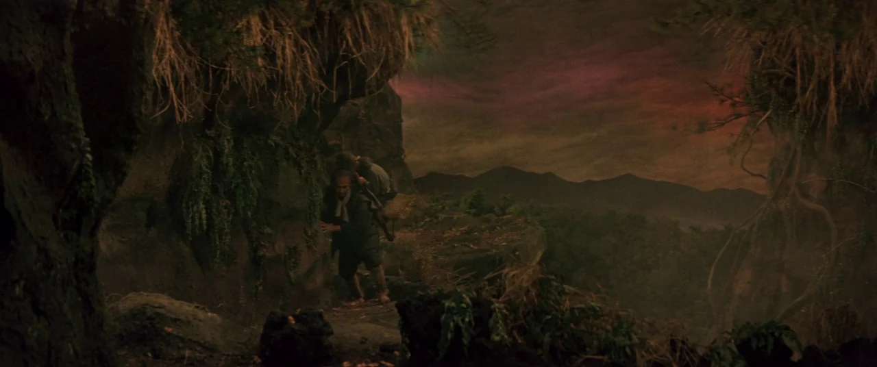 Ballad Of Narayama (1958) movie still: wide — A dark, craggy mountain trail winds along the edge of a steep precipice. A man carries a smaller…