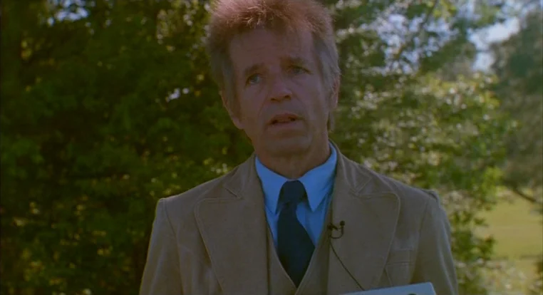 Schizopolis movie still: medium — An outdoor park setting with blurry green trees visible in the background. A middle-aged man in a suit speaks…