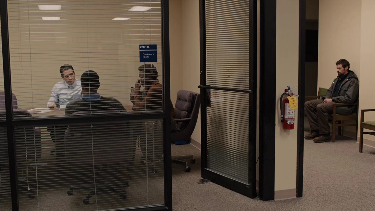 Prisoners (2013) movie still: wide — An administrative area with a glass-enclosed meeting room beside a beige hallway wall. A man sits alone in the…