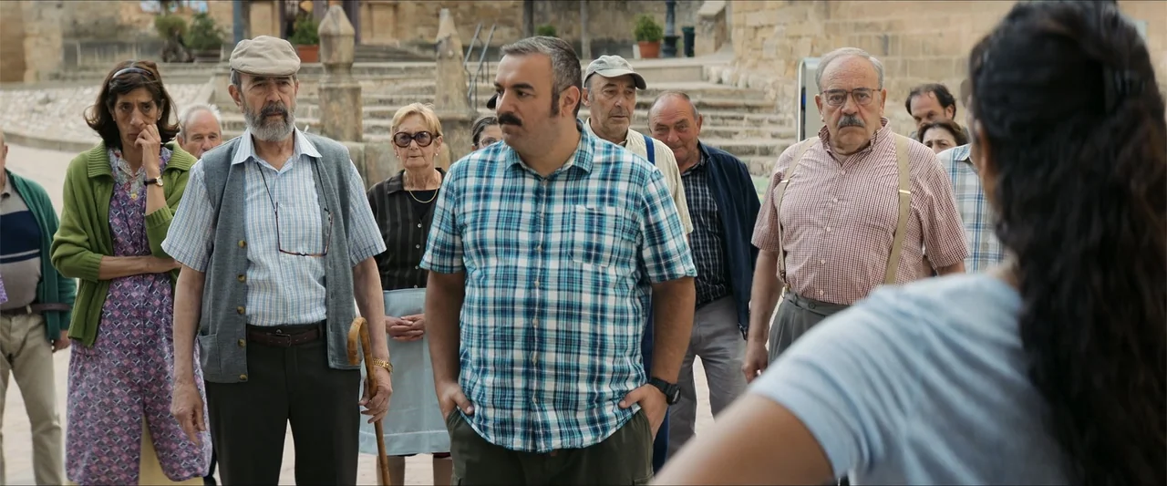 A Hipster in Rural Spain (2024) movie still: medium — A group of older adults gathers in a stone-paved European style town square.