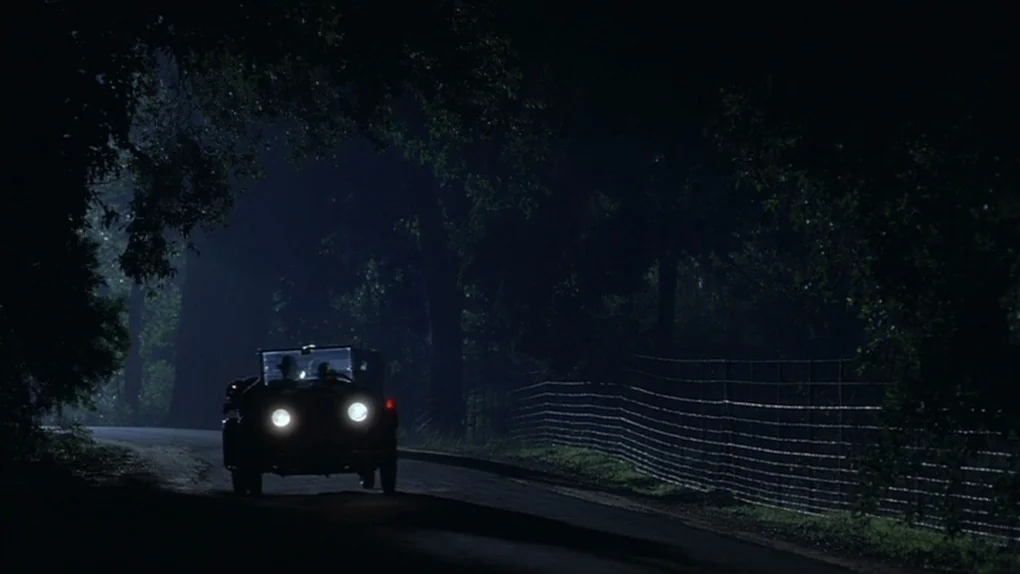 Ask The Dust (2006) movie still: wide — A dark road winds through a heavily wooded area at night. A vintage automobile with glowing headlights moves…