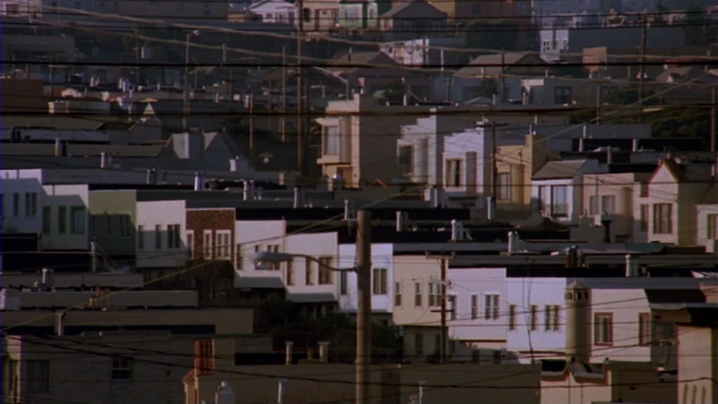 Patty Hearst (1988) movie still: establishing — A dense cluster of suburban residential buildings extends across a hillside cityscape. Numerous…
