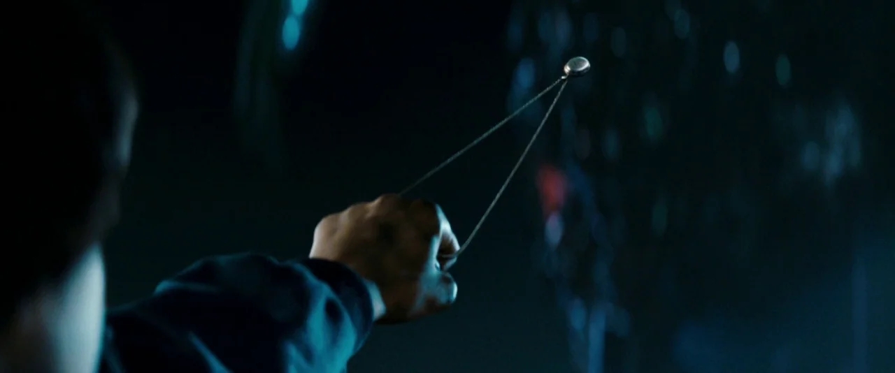 Super 8 (2011) movie still: closeup — A dark environment obscures most details of the surroundings. A hand holds a silver pendant suspended by a…