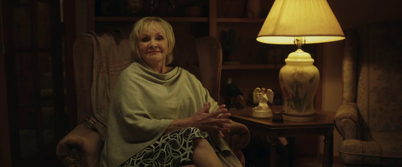 LaRoy, Texas (2023) movie still: medium — An older woman sits in an armchair inside a dim room with bookshelf backdrops. She has her hands clasped…