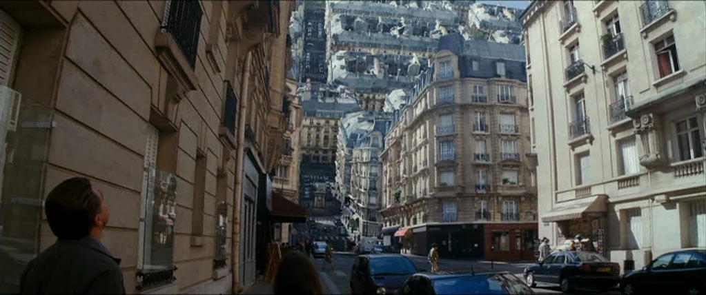 Inception (2010) movie still: wide — A Parisian street with classic Haussmann buildings folds upward into the sky. A man stands in the foreground…