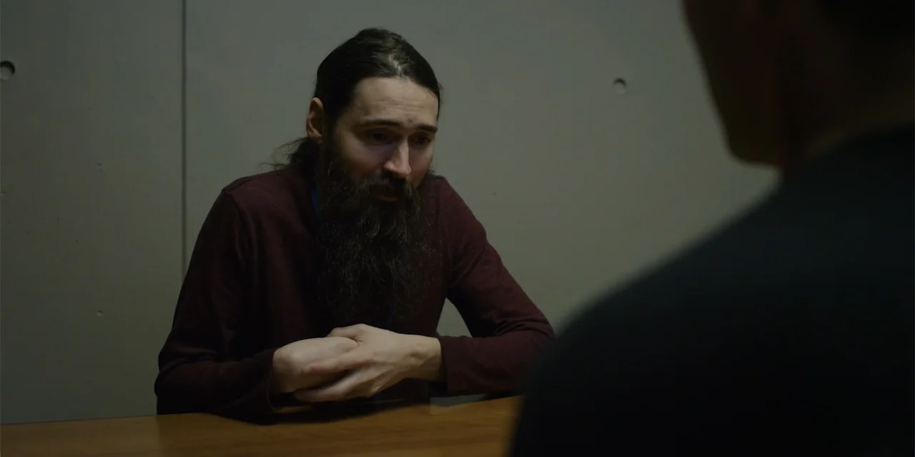 Lobos (2019) movie still: medium — An interrogation room features a plain gray wall with visible concrete construction joints. A bearded man in a…