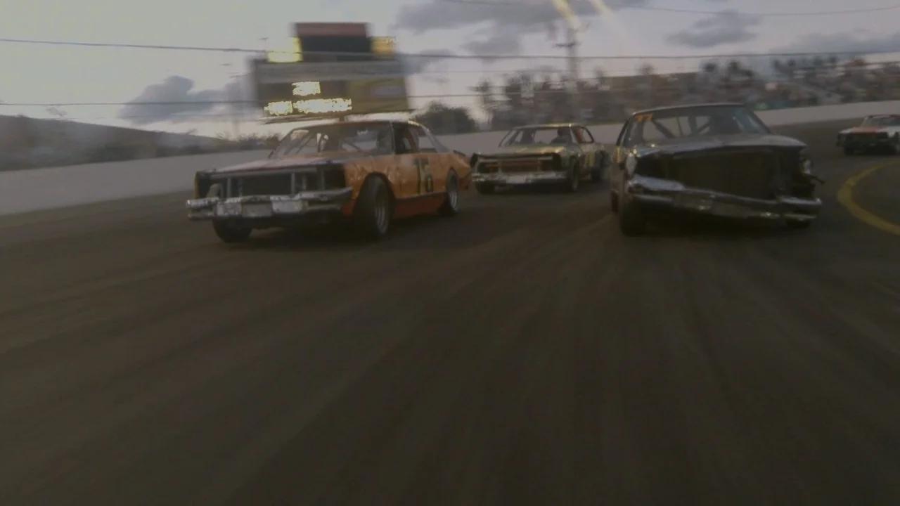 The Last Race (2022) movie still: wide — A group of damaged stock cars race around a paved oval track at dusk. The cars navigate a sharp turn while…