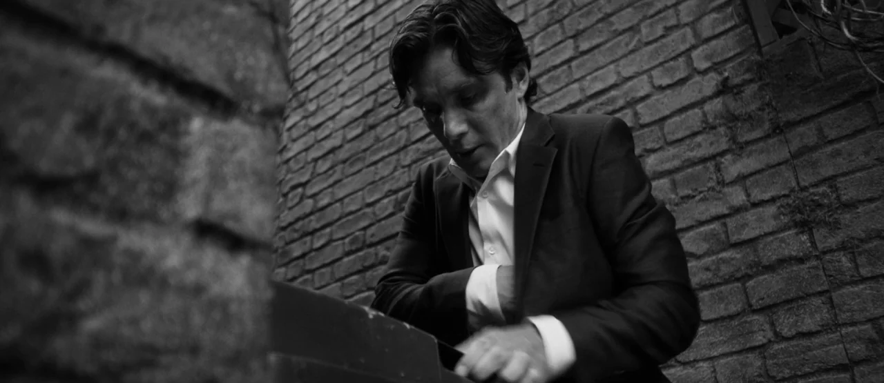 The Party (2017) movie still: medium — A man stands before a rough textured brick wall in an narrow outdoor space. He wears a dark suit and reaches…