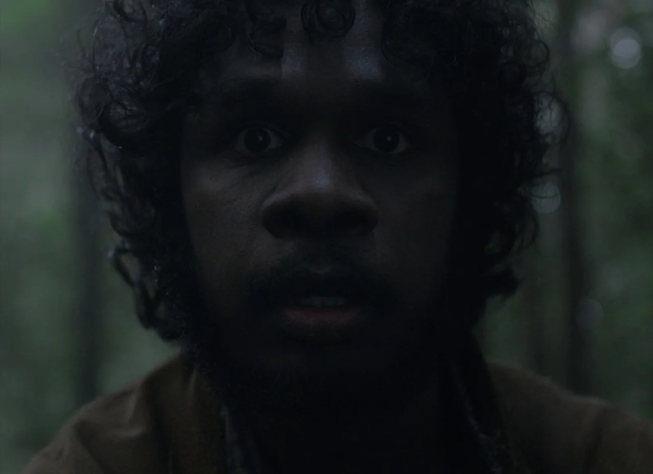 The Nightingale (2018) movie still: closeup — A dense, dimly lit forest background with soft out-of-focus greenery. A young man faces forward with a…