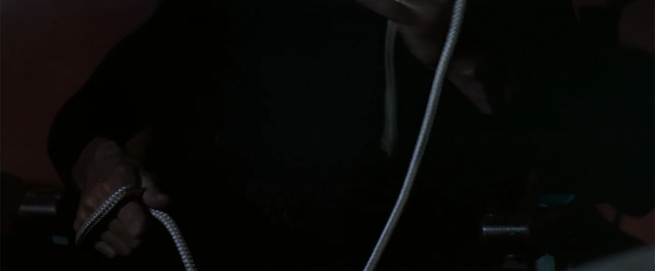 The Pelican Brief (1993) movie still: closeup — A dark interior space features a white cord being manipulated by a hand. The rope hangs vertically…