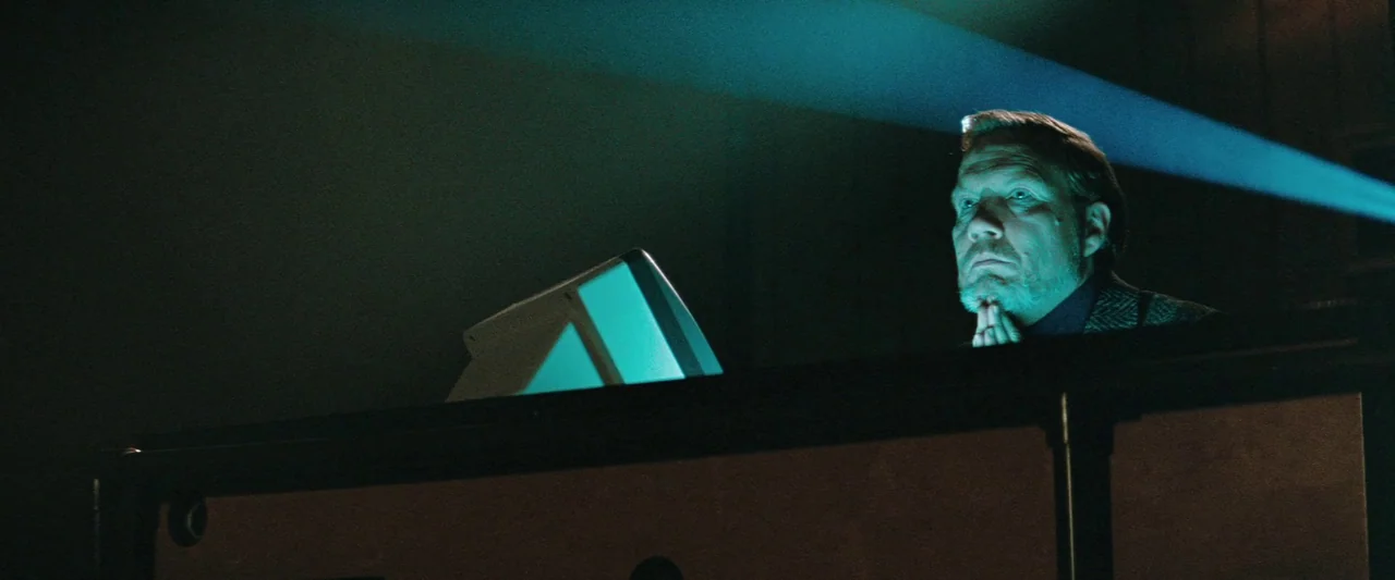The Adjuster (1991) movie still: medium — A darkened interior room with a metal railing and a glowing projector screen. An older man sits behind the…