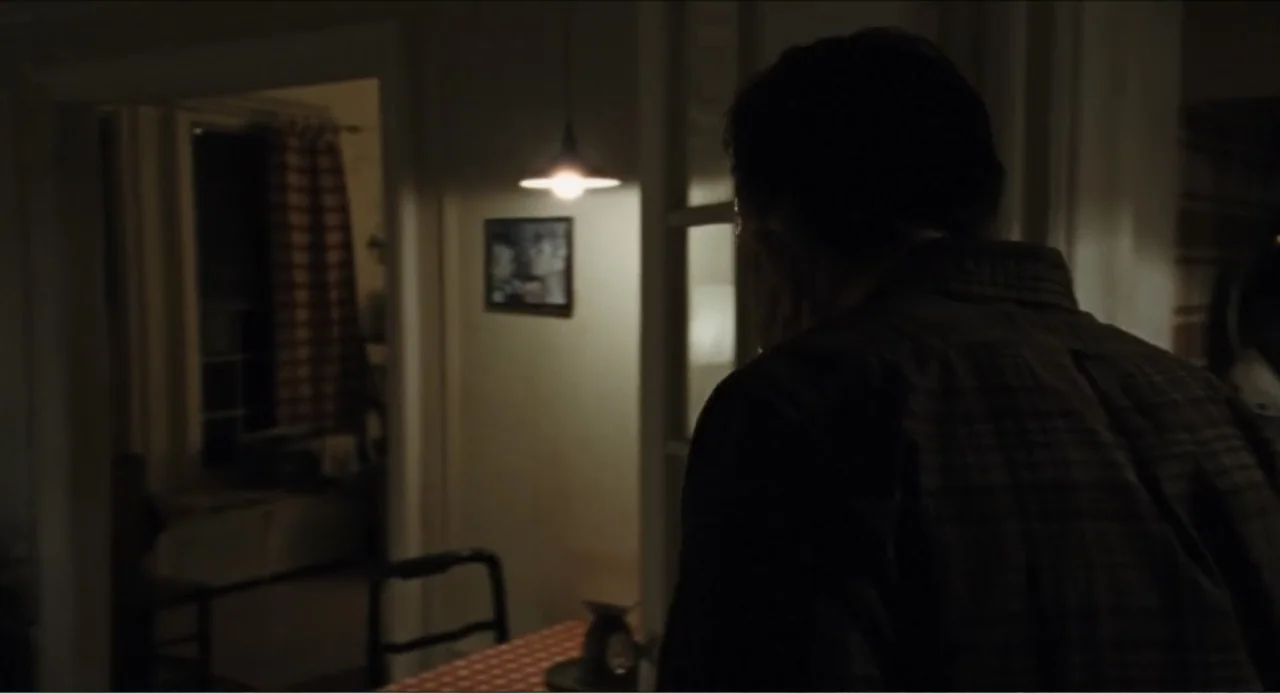 A Different Man (2024) movie still: medium — A dimly lit room with a hanging lamp over a patterned tablecloth in distance. A man in a dark plaid…