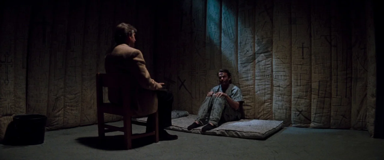 In The Mouth of Madness (1994) movie still: wide — A dimly lit room features padded walls covered in hand-drawn cross symbols. A man sits on a floor…