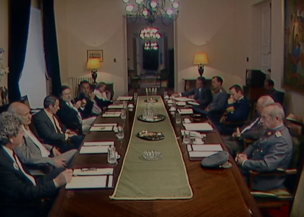 No (2012) movie still: wide — A formal meeting room features a long wooden table with twelve men seated around. Papers and glasses are arranged…