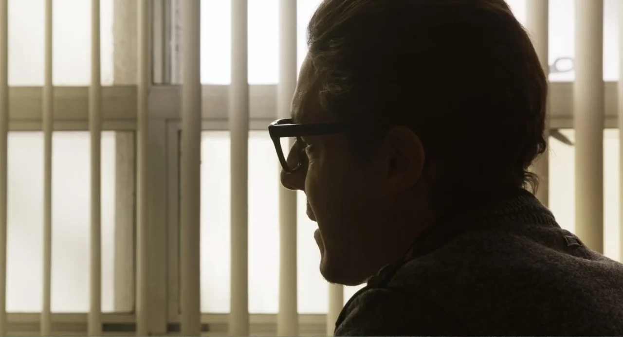 Salvation (2025) movie still: closeup — A man sits in front of a window with vertical blinds covering the glass. The subject is in partial…