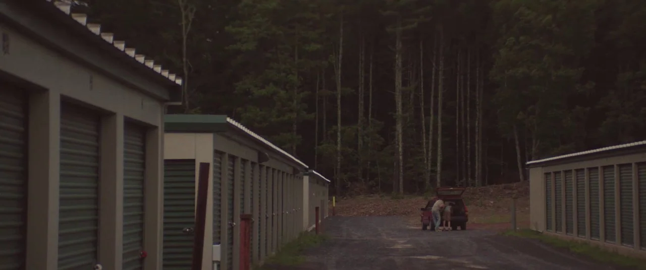 The Strange Ones (2017) movie still: wide — A gravel driveway passes between lines of beige storage units with green roll-up doors. Two figures…