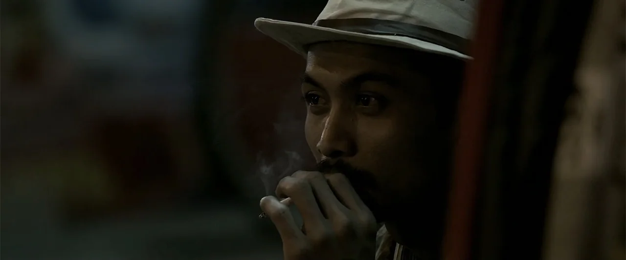 The Kleptocrats (2018) movie still: closeup — A dark outdoor urban setting at night features a man in a fedora. The man smokes a cigarette while…
