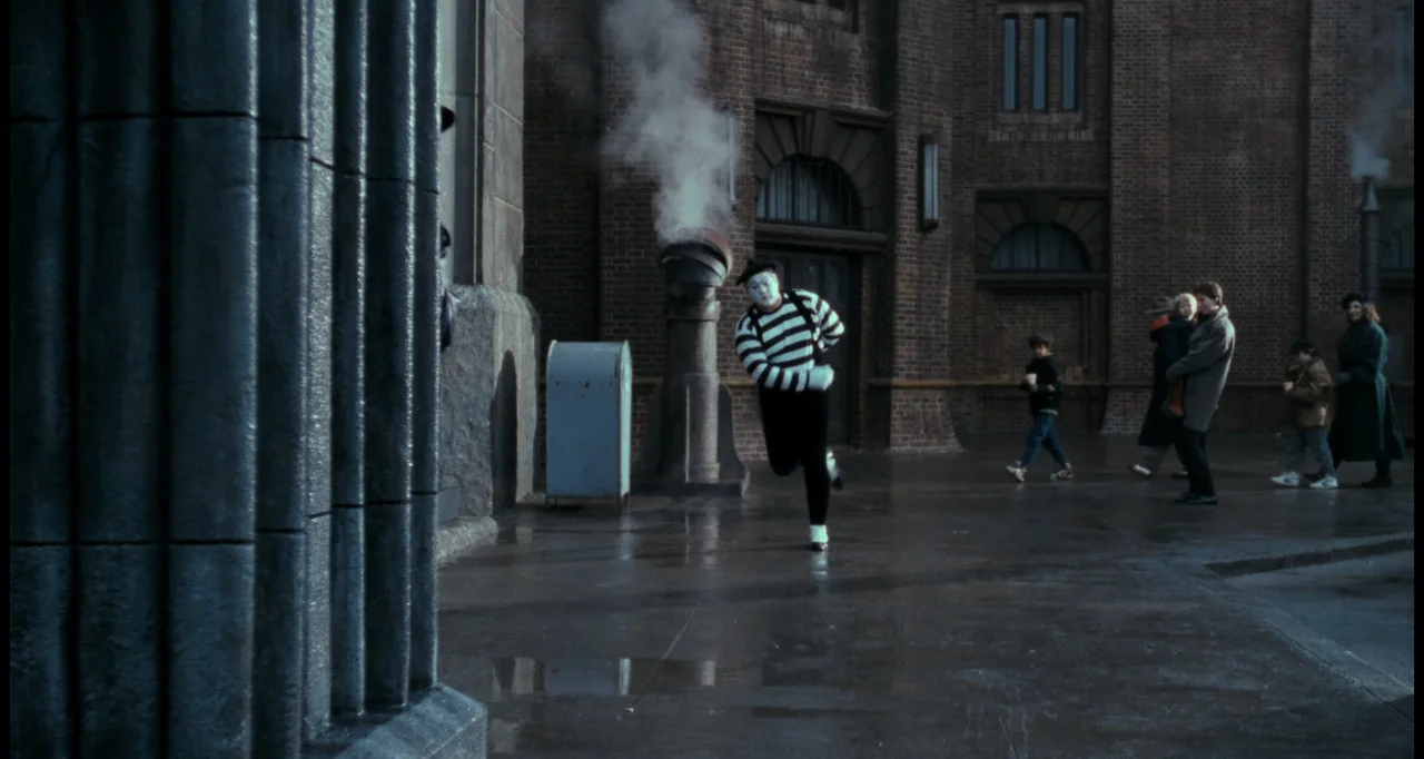 Batman (1989) movie still: fullbody — A brick-walled city courtyard features wet pavement and a steaming metal vent pipe. A mime wearing a striped…