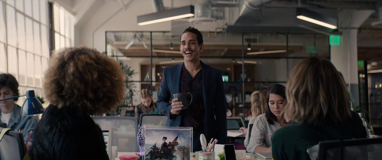 Tone Deaf (2019) movie still: medium — A sunlit modern open-plan office features glass walls and rows of workstations. A man holding a coffee mug…