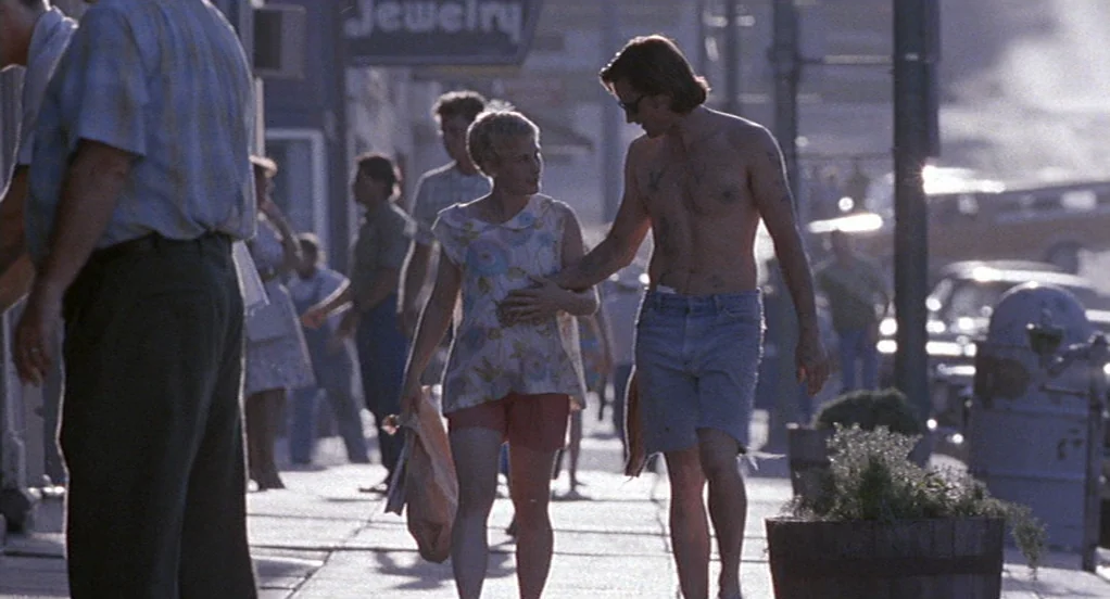 The Indian Runner (1991) movie still: fullbody — A sunny pedestrian walkway lined with cars and distant storefronts under a clear sky. A young…