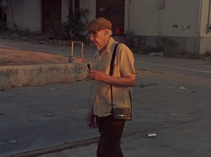 Slacker (1991) movie still: fullbody — An elderly man walks through a desolate, paved industrial lot during sunset. He holds a small wired…