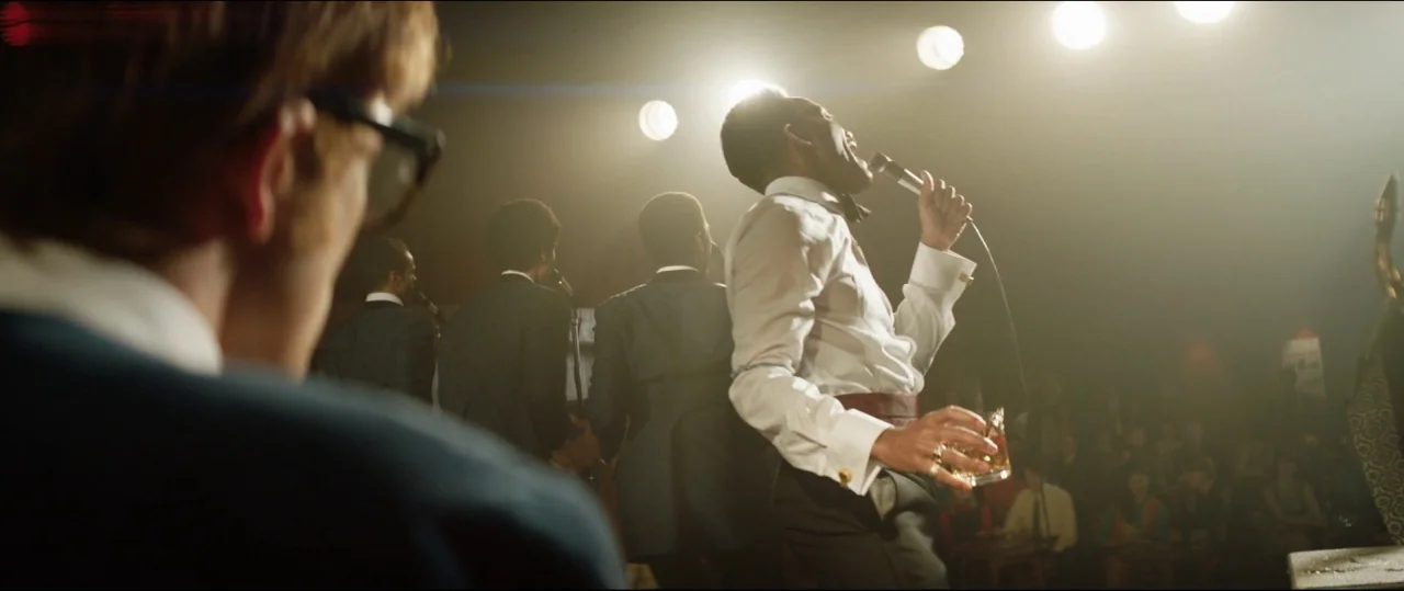 Rocketman (2019) movie still: medium — A dimly lit club stage features a singer and band members in dark suits. The singer performs into a…