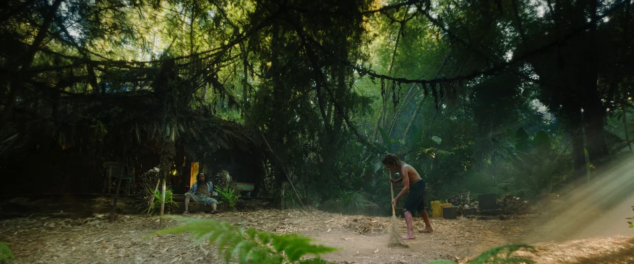 Boy Kills World (2023) movie still: wide — A clearing within a dense jungle featuring a rustic shelter constructed of natural materials. A man…