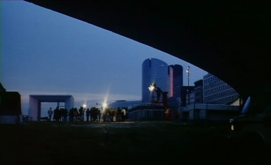 Simon, The Magician (1999) movie still: wide — A dark, grassy vacant lot is framed by a heavy diagonal architectural shadow. A group of people…