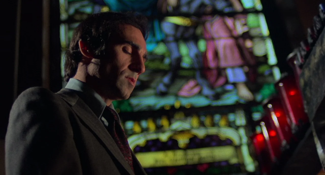 God Told Me To (1976) movie still: closeup — A man stands in a dark interior space against a colorful, lit stained glass window. The stained glass…