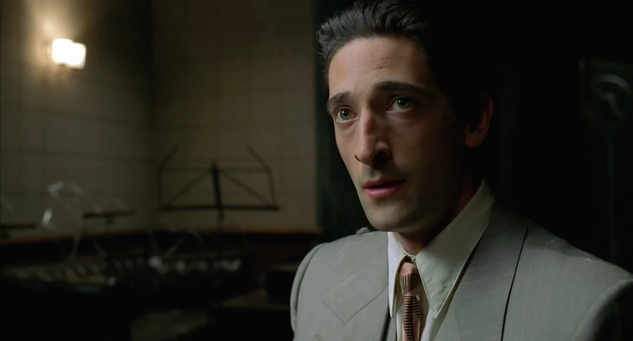 The Pianist (2002) movie still: closeup — A dimly lit rehearsal space features wall-mounted fixtures and blurred music stands in the background. A…