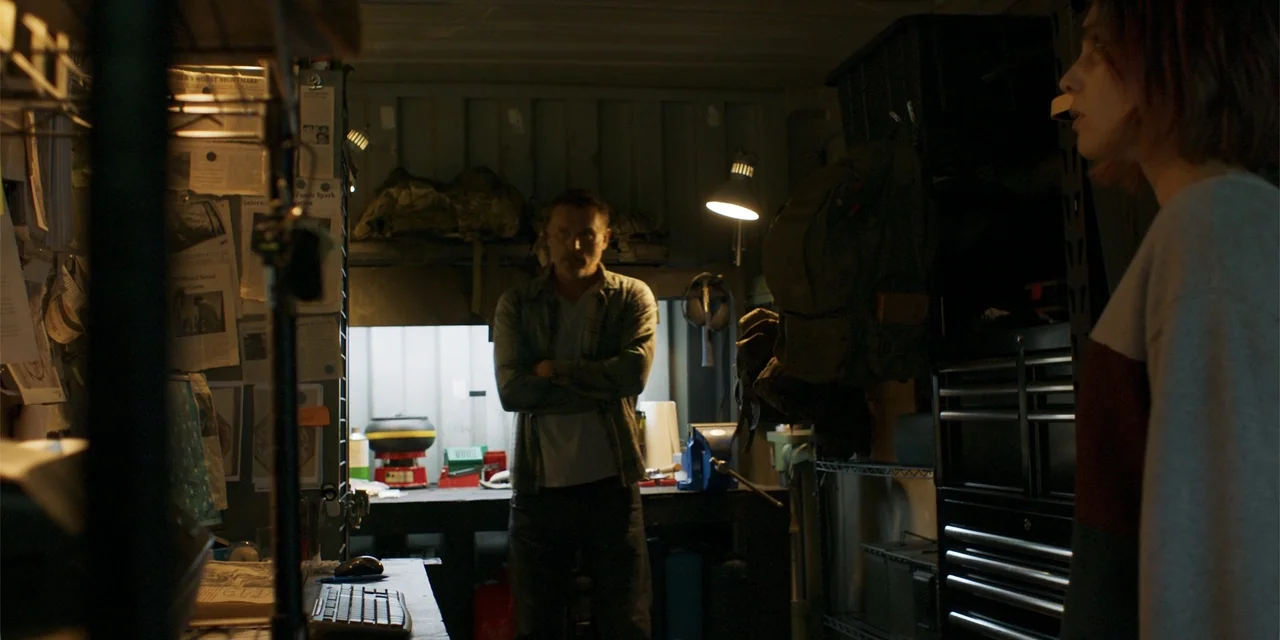 Demonic (2015) movie still: medium — A cluttered, dimly lit storage container serves as a makeshift workspace with walls covered in documents. A man…