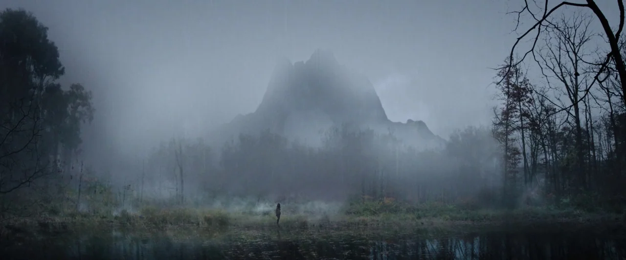 Wildling (2018) movie still: wide — A dense foggy swamp with a large dark rock formation looming in the distance. A lone figure stands motionless in…