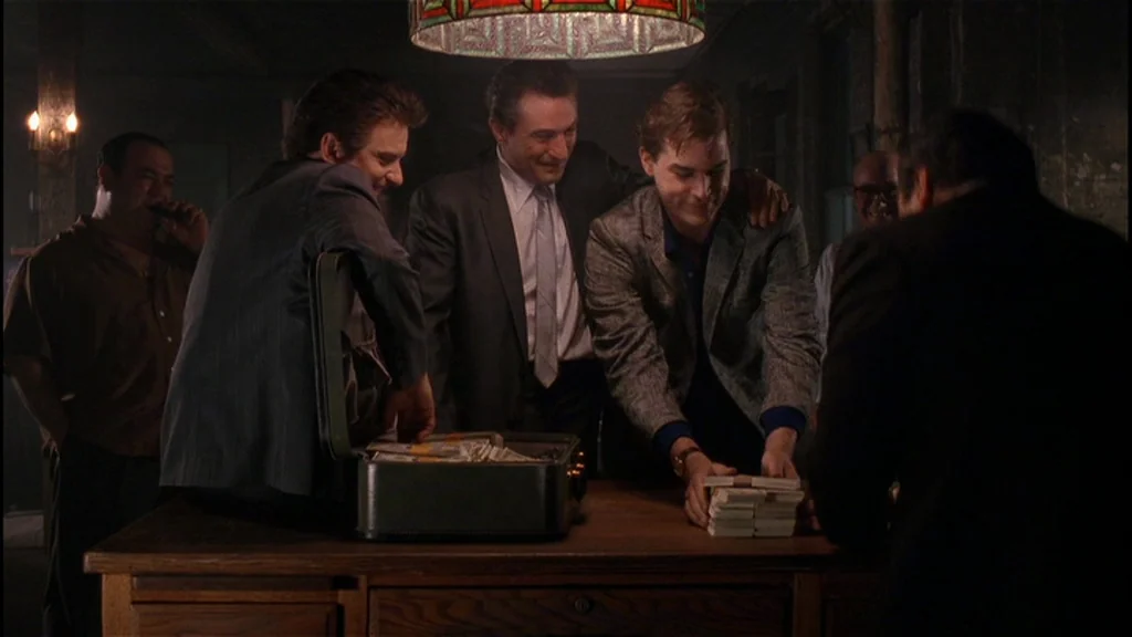 Goodfellas (1990) movie still: medium — A dark, wood-paneled room is occupied by a group of men gathered closely. They are huddled around a desk…