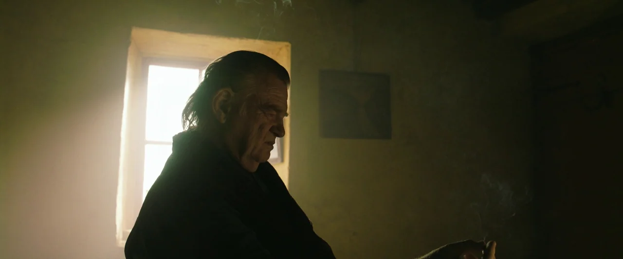 The Banshees of Inisherin (2022) movie still: medium — A dark, dimly lit room with plain walls features light pouring through a window. An older man…