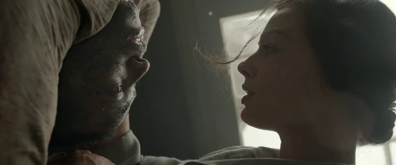 Amsterdam (2022) movie still: closeup — An interior space features two people in close proximity in front of a window. A woman faces a man whose…