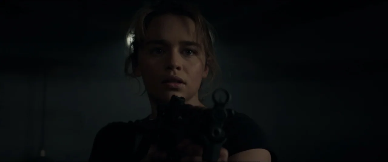 Terminator Genisys (2015) movie still: closeup — A dimly lit room with heavy shadows obscuring architectural details. A woman in a black shirt holds…
