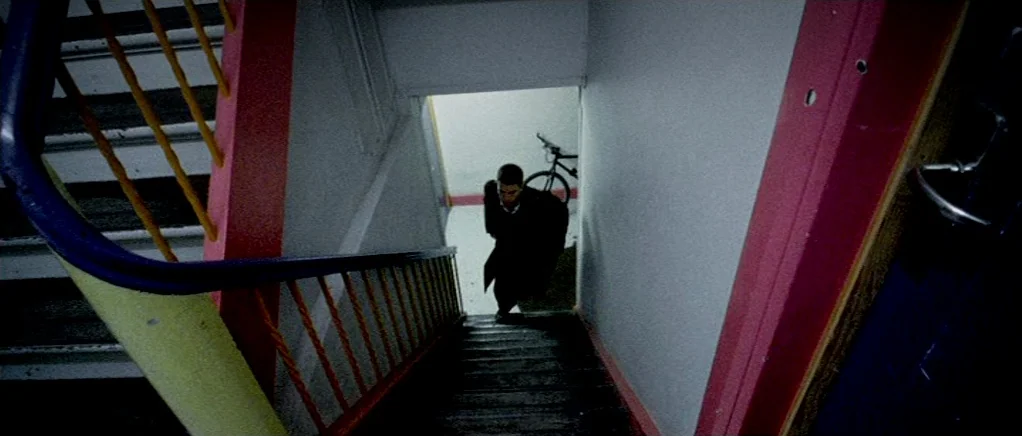 Reconstruction (2003) movie still: wide — A stairwell features bright red and yellow painted metal railings against white walls. A young man in a…