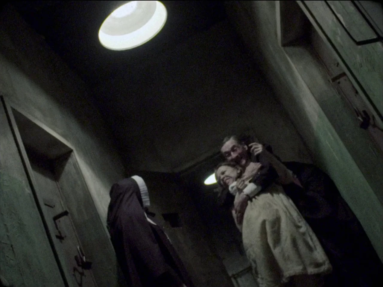 The Devil’s Doorway (2018) movie still: medium — A dimly lit concrete hospital corridor with heavy metal doors and overhead circular lamps. A nun…