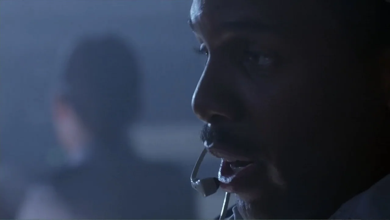 Crisis Point (2012) movie still: closeup — A dimly lit room with blue ambient light fills the background space. A man wearing a headset speaks while…